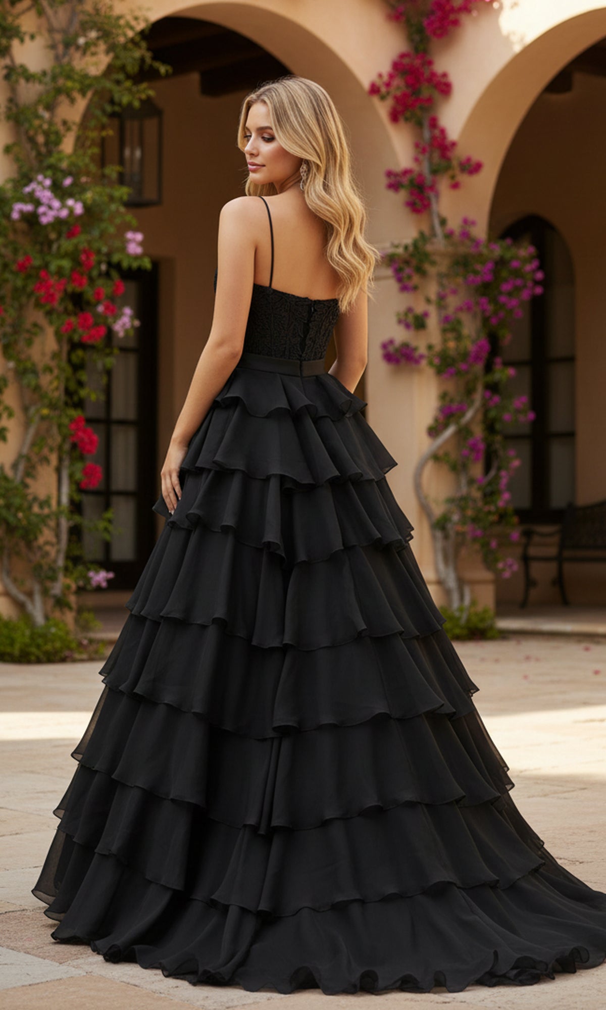 A woman wearing the Long Prom Dress: PromGirl Exclusive 250202 stands outdoors, slightly turned away. Bougainvillea vines climb a beige building behind her.